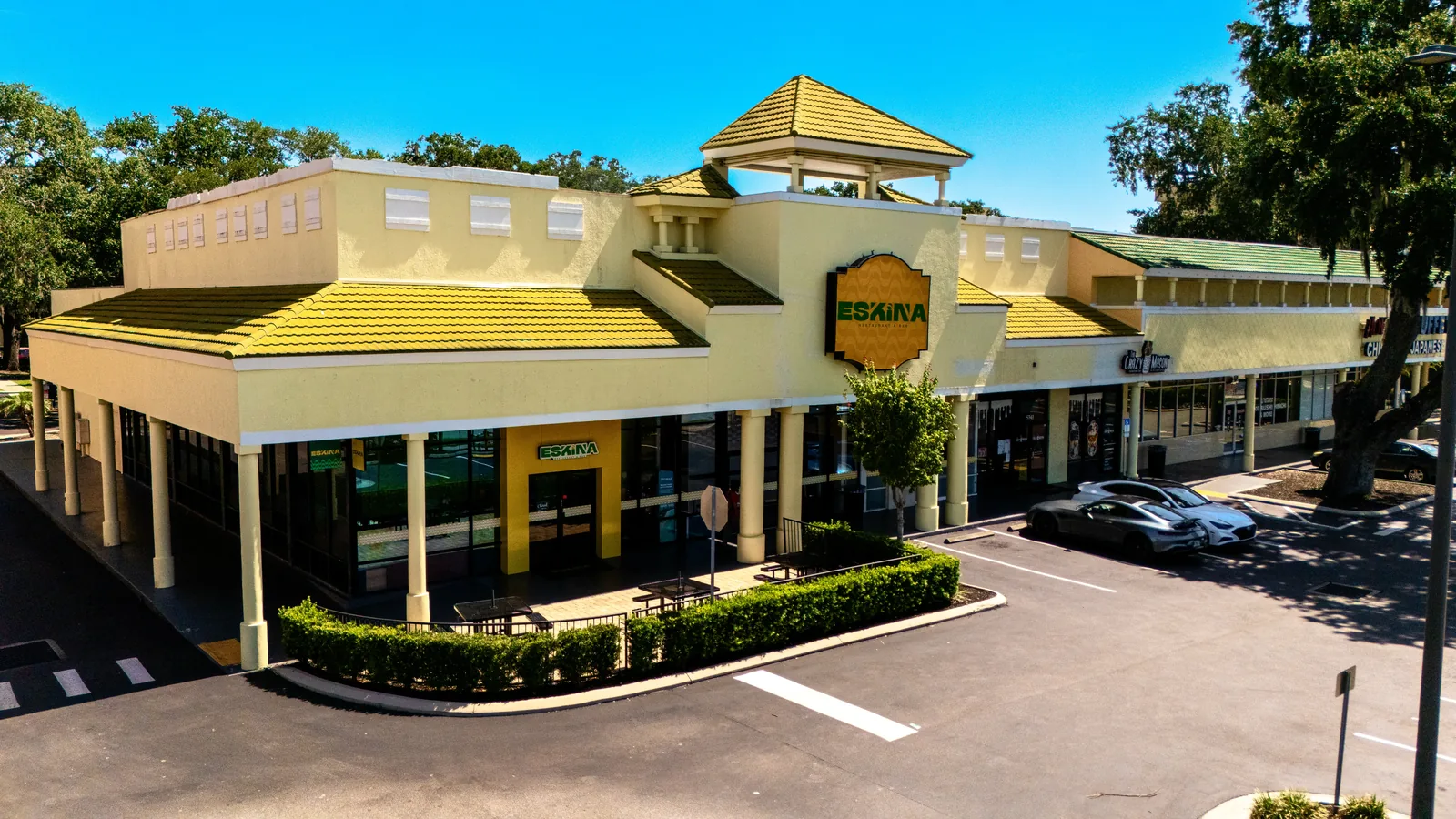 Aerial view of an Eskina restaurant storefront in a gray shopping center, featuring large Eskina signage, black awnings, surrounding storefronts, trees, and an empty parking area.