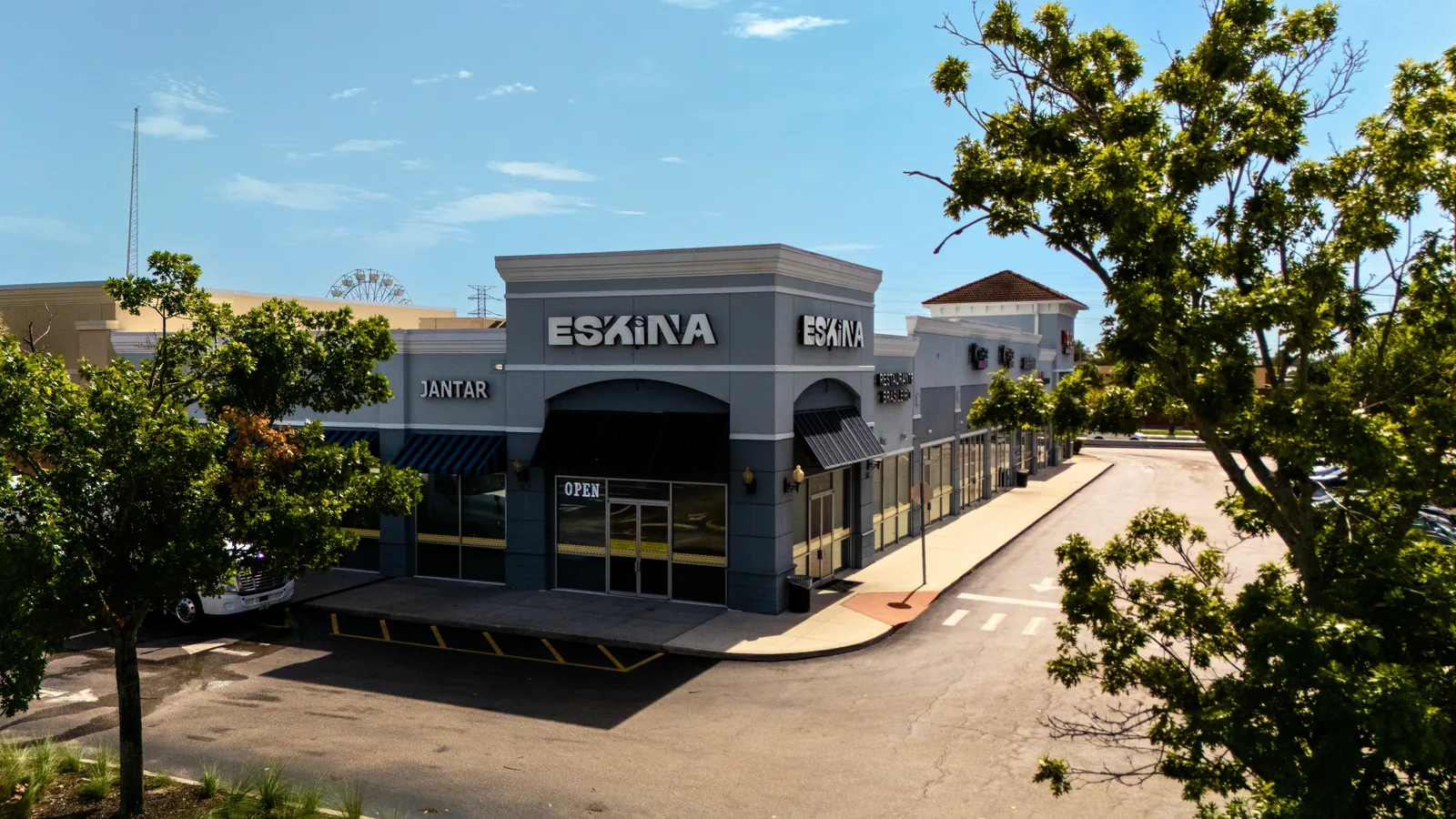 Aerial view of an Eskina restaurant storefront in a gray shopping center, featuring large Eskina signage, black awnings, surrounding storefronts, trees, and an empty parking area.